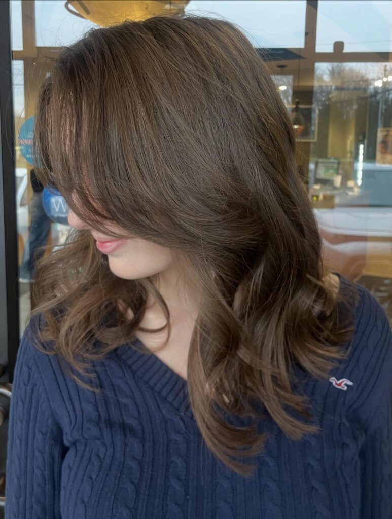 A woman with medium length glossy mocha brown hair featuring soft layers and sweeping curtain bangs with curled ends.