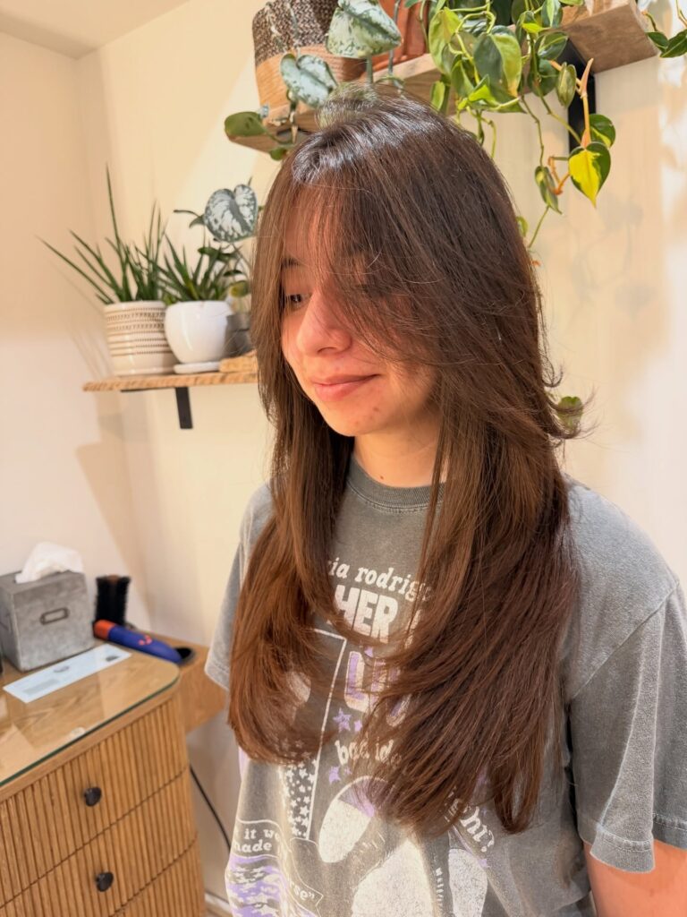 A woman with long layered chestnut brown hair and wispy bangs framing her face.
