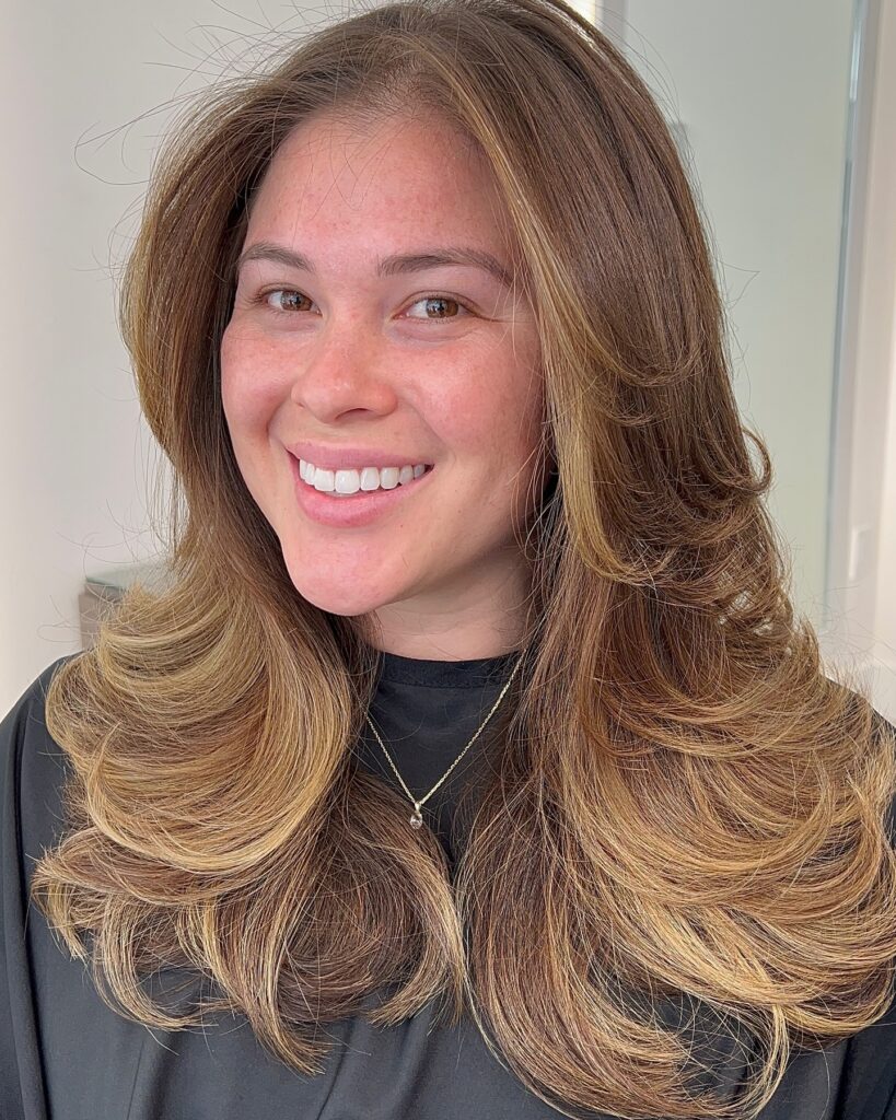 A smiling woman with long layered brown hair featuring voluminous caramel highlights and soft outward-curling ends.