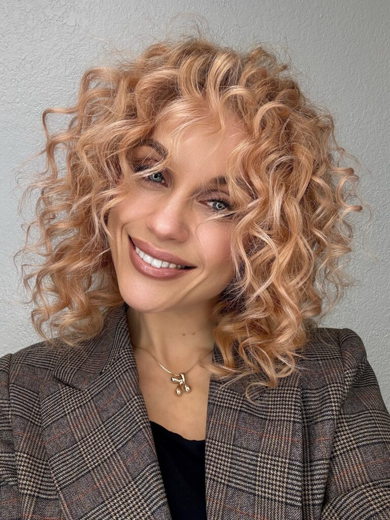 A woman with a voluminous blonde curly bob featuring soft layers and defined ringlets.