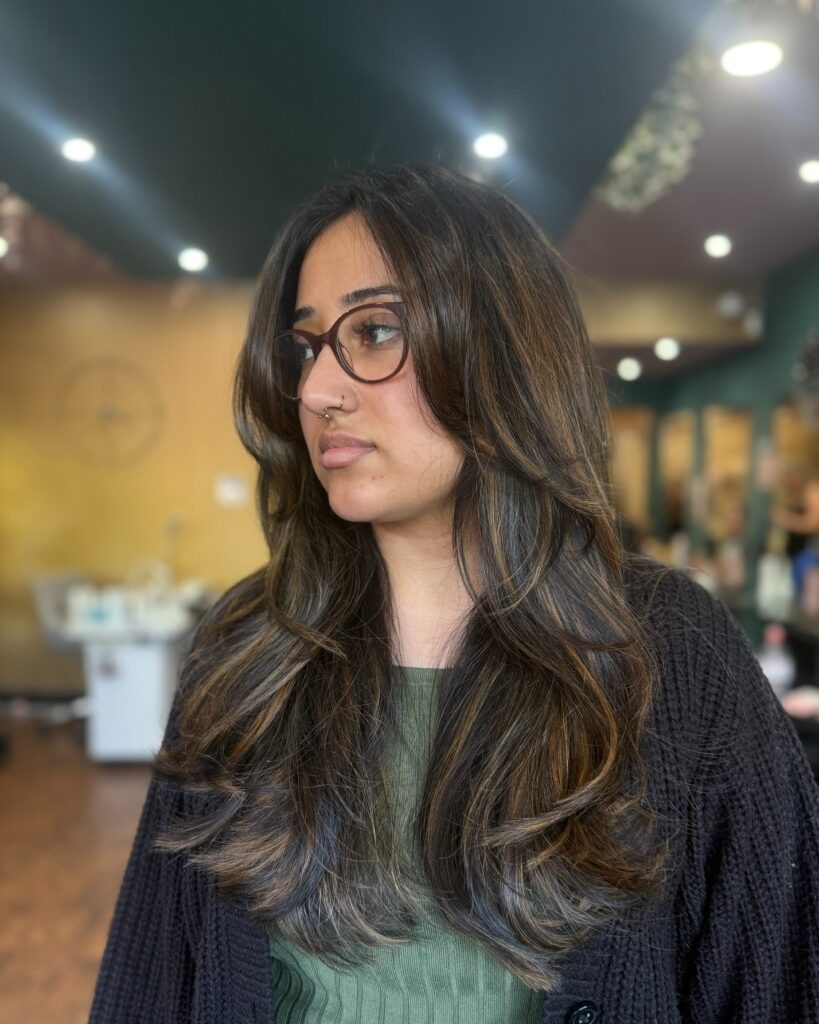 A woman with long wavy brunette hair with caramel brown highlights and face framing butterfly layers.