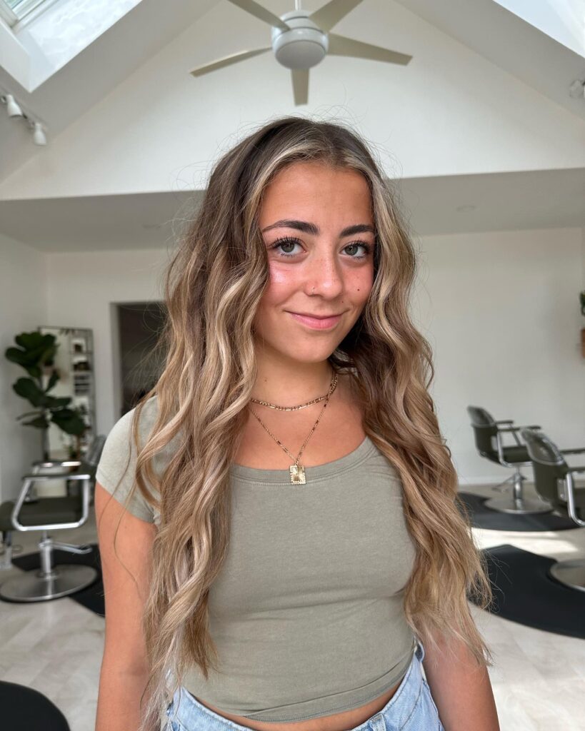 A smiling woman with long bronde hair in wavy balayage with prominent light blonde face-framing streaks.