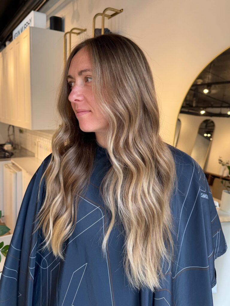 A woman with very long wavy hair featuring a sandy blonde balayage and a soft side profile view.