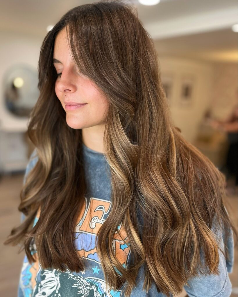 A woman with long brunette hair featuring warm caramel balayage highlights and soft beachy waves.