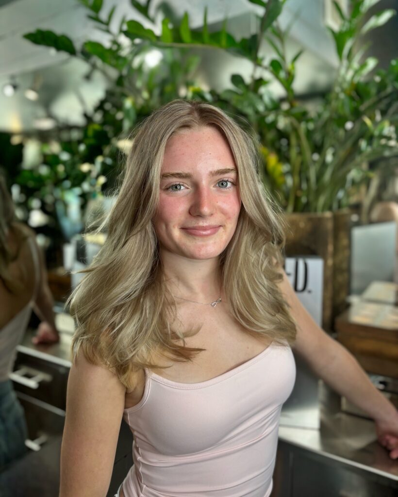 A smiling woman with mid length creamy blonde hair featuring soft face framing layers and a voluminous texture.