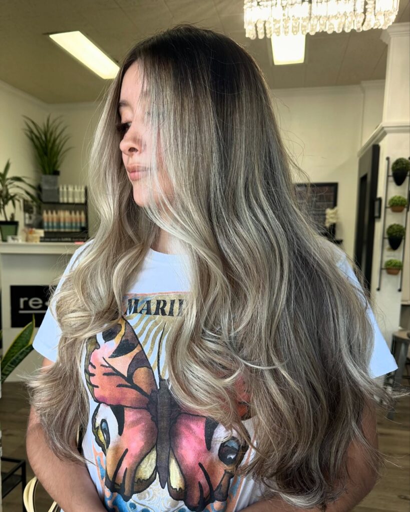 A woman with very long, thick ash blonde balayage hair featuring prominent money piece highlights and voluminous waves.