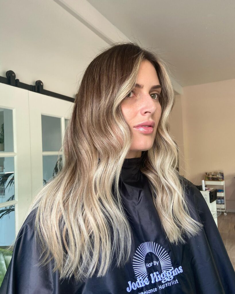 A woman with long honey blonde hair featuring soft butterfly layers and long curtain bangs.