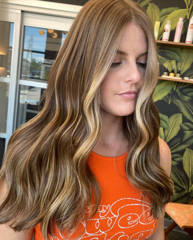 A woman with long wavy brown hair featuring warm honey blonde balayage and bright face framing highlights.