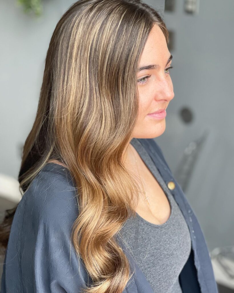 A woman with long wavy brown hair featuring prominent caramel balayage highlights styled over her shoulder.