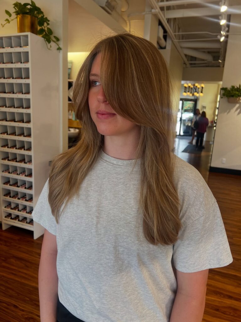 A woman with long honey blonde hair featuring soft butterfly layers and face framing curtain bangs.