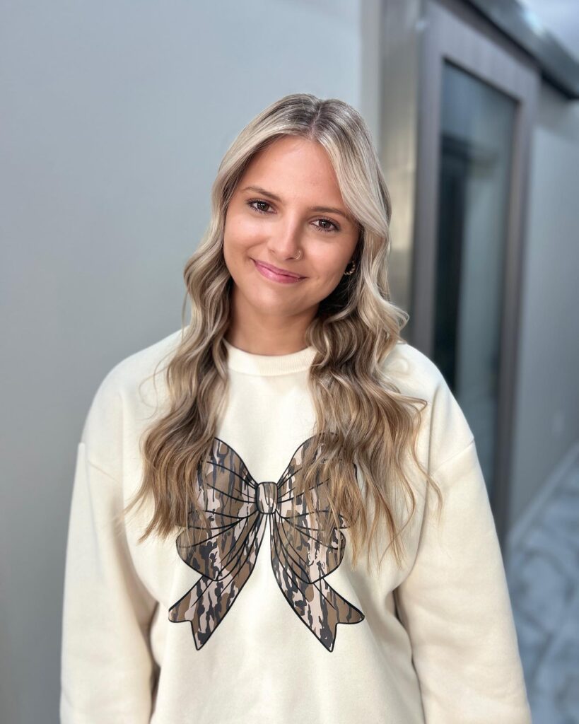 A smiling woman with long wavy blonde hair featuring bright face framing highlights and soft textured layers.