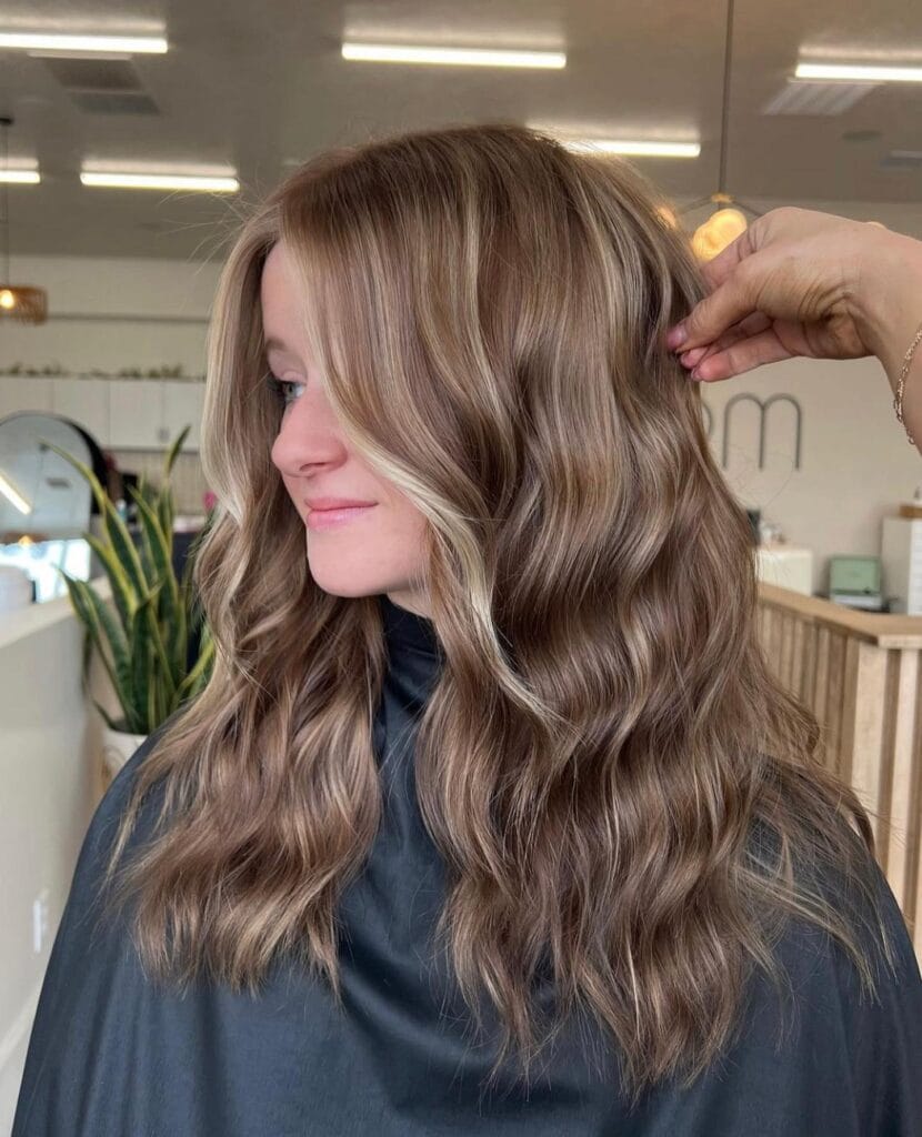 A woman with long wavy dark blonde hair featuring bright face framing money piece highlights and a middle part.