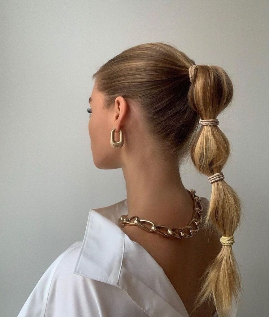 A woman with blonde hair styled in a high bubble ponytail with gold hair ties and chunky gold jewelry.