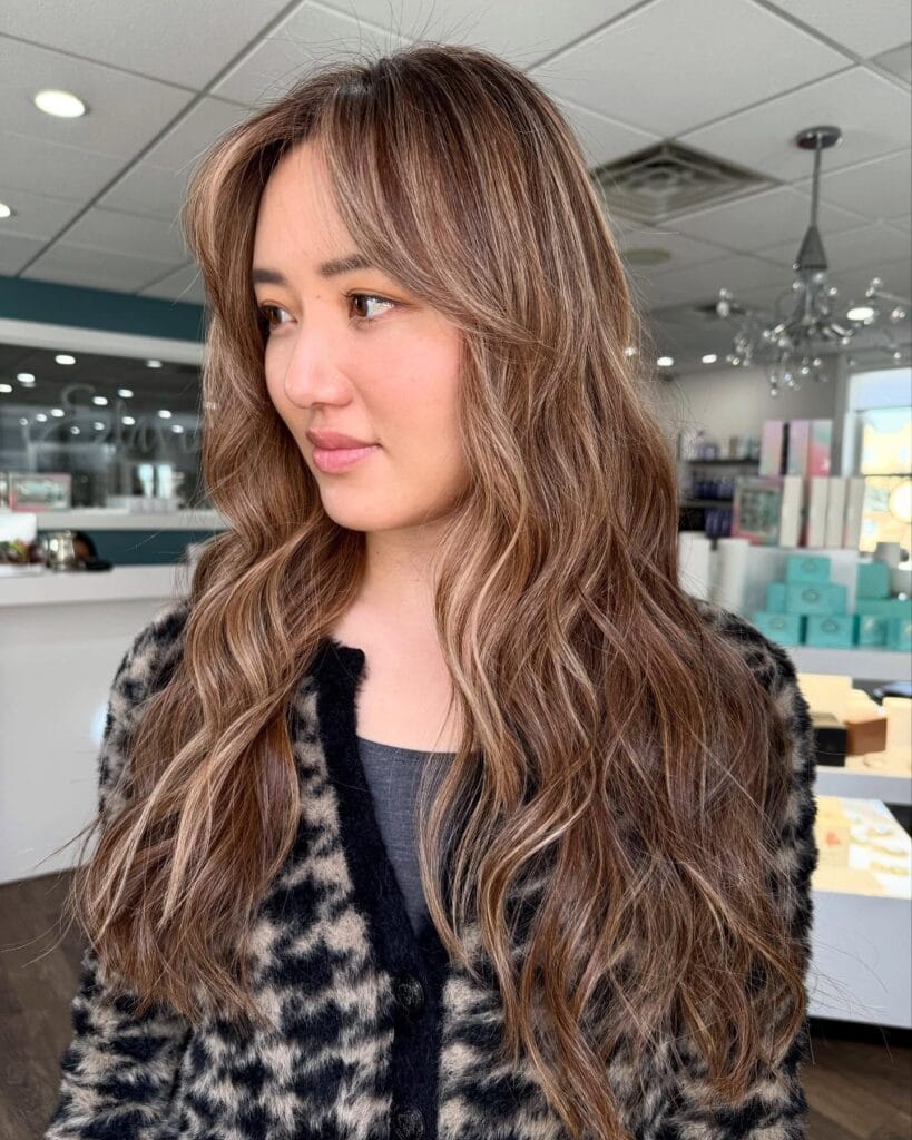 A three-quarter profile shot of a woman with long thick brunette hair featuring face framing layers and soft curled ends, styled with bright money piece highlights.