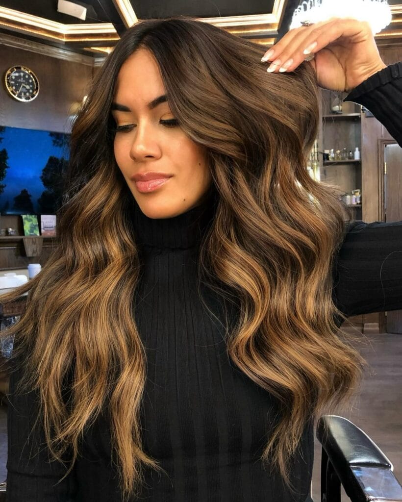 A woman with very long, thick, dark hair and honey blonde balayage, styled in large loose waves and parted in the center.