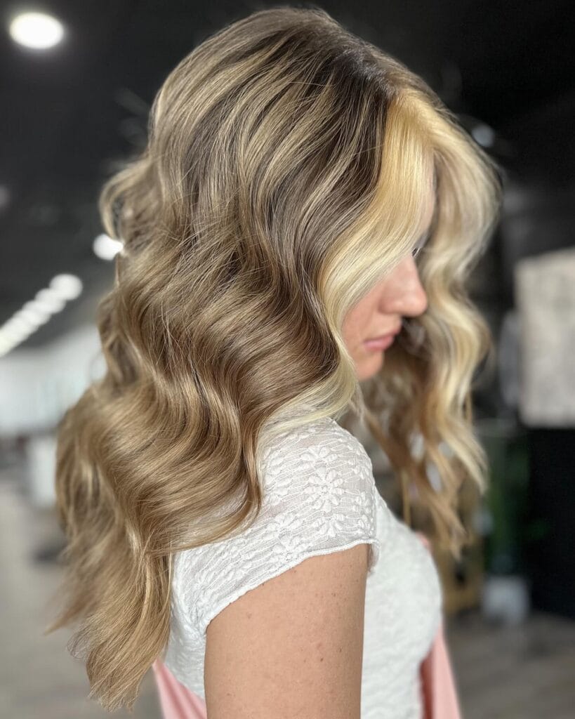 A side profile view of a woman with long thick ash blonde hair styled in large glossy waves featuring bright face framing highlights.