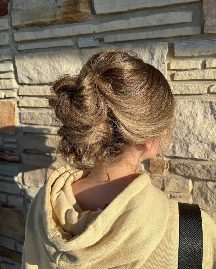 A woman with honey blonde wavy hair wears a voluminous messy low bun with soft curled strands framing her face.