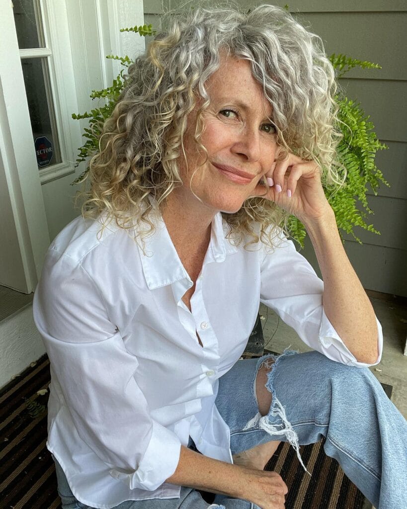 A woman with shoulder-length voluminous silver and blonde curly hair wearing a white shirt and ripped jeans.