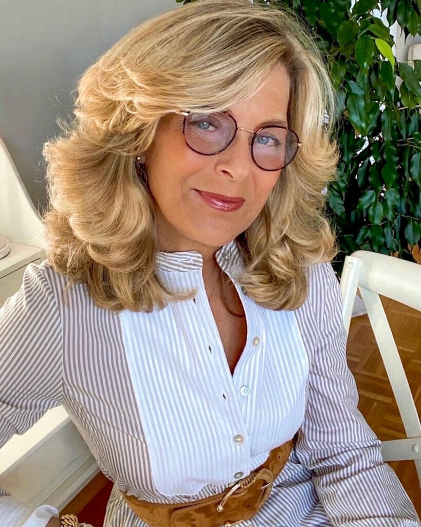 A woman with voluminous shoulder-length golden blonde hair styled in feathered layers and soft curls.