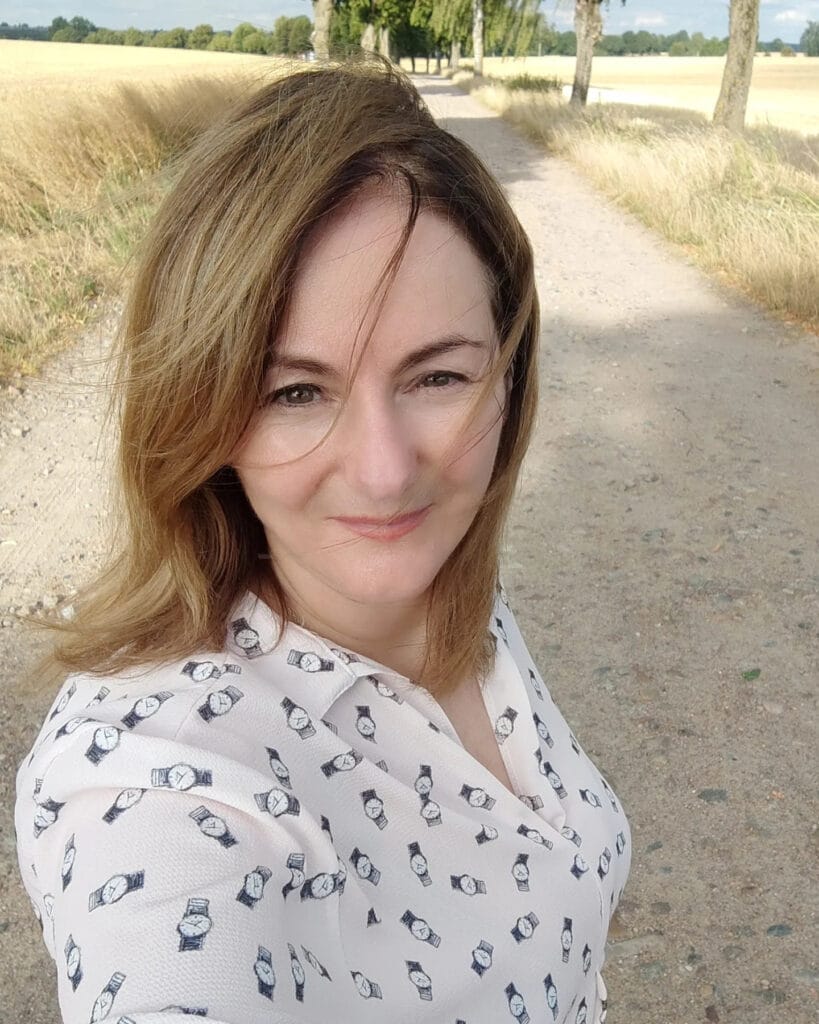 A woman with shoulder-length warm honey-blonde hair styled in a natural wind-swept lob is standing outside on a dirt path.
