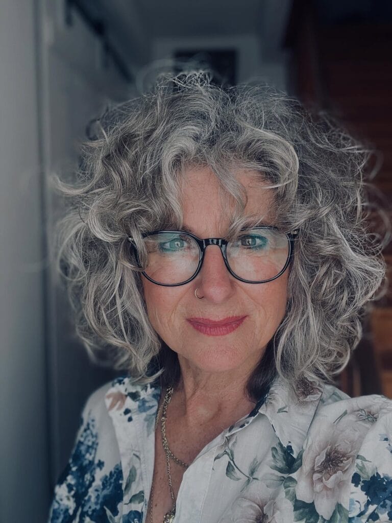 A woman with voluminous shoulder-length salt and pepper curly hair wearing black rimmed glasses and a floral shirt.