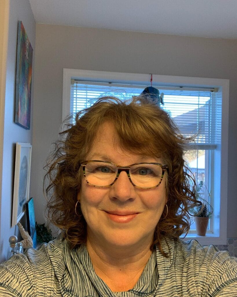 A woman with shoulder-length messy brown curls and glasses is smiling for a photo.