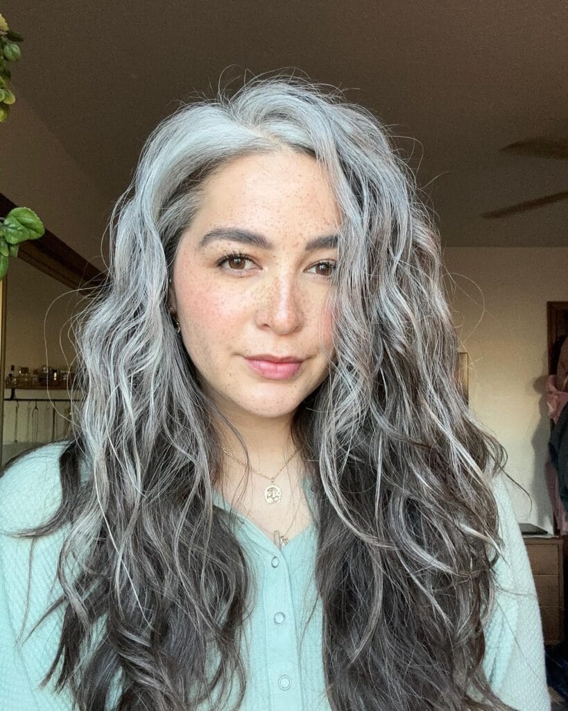 A woman with long, cascading salt-and-pepper hair styled in loose, beachy waves.