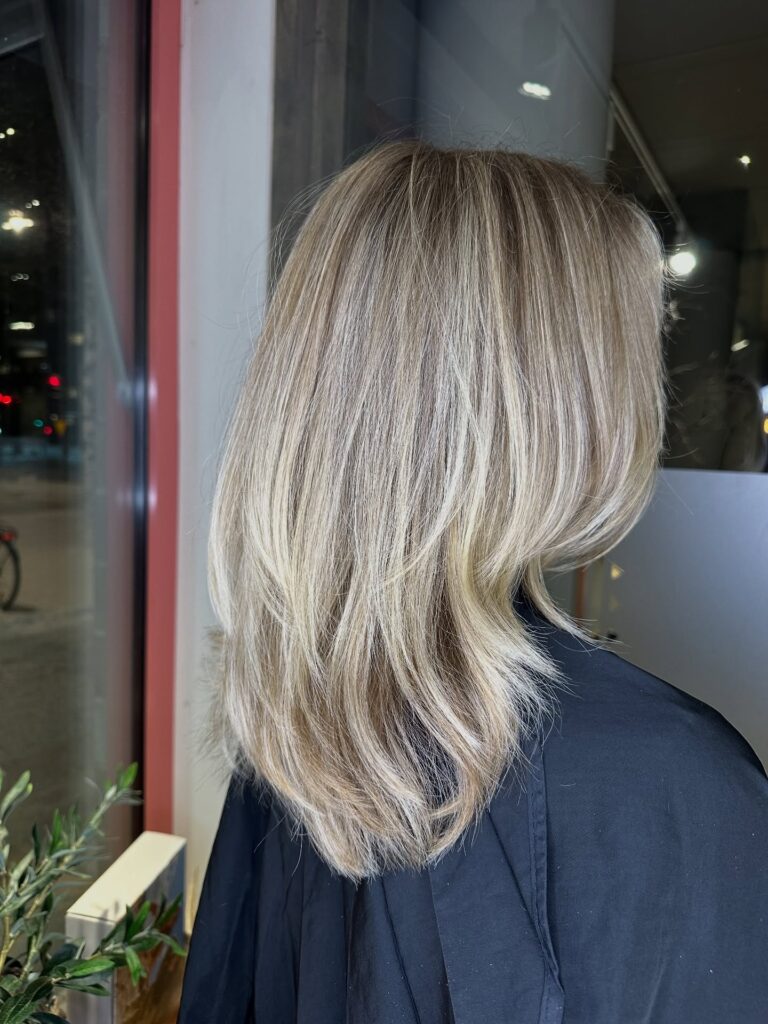 A woman from the back with medium-length ashy blonde hair featuring soft, feathered layers and bright highlights.