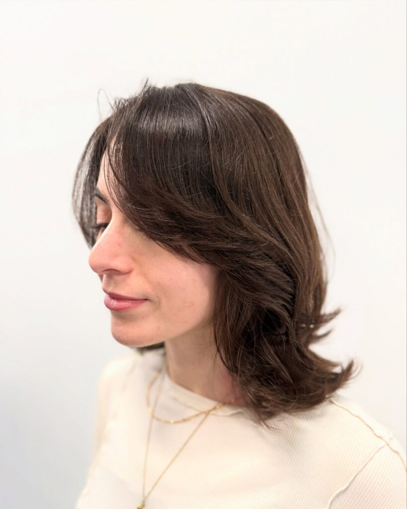 A woman from the side with a medium-length chestnut brown haircut featuring soft layers and a side-swept fringe.