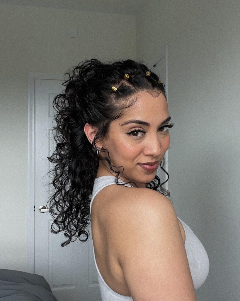 A woman with medium-length black curly hair tied in a high ponytail decorated with gold hair cuffs.