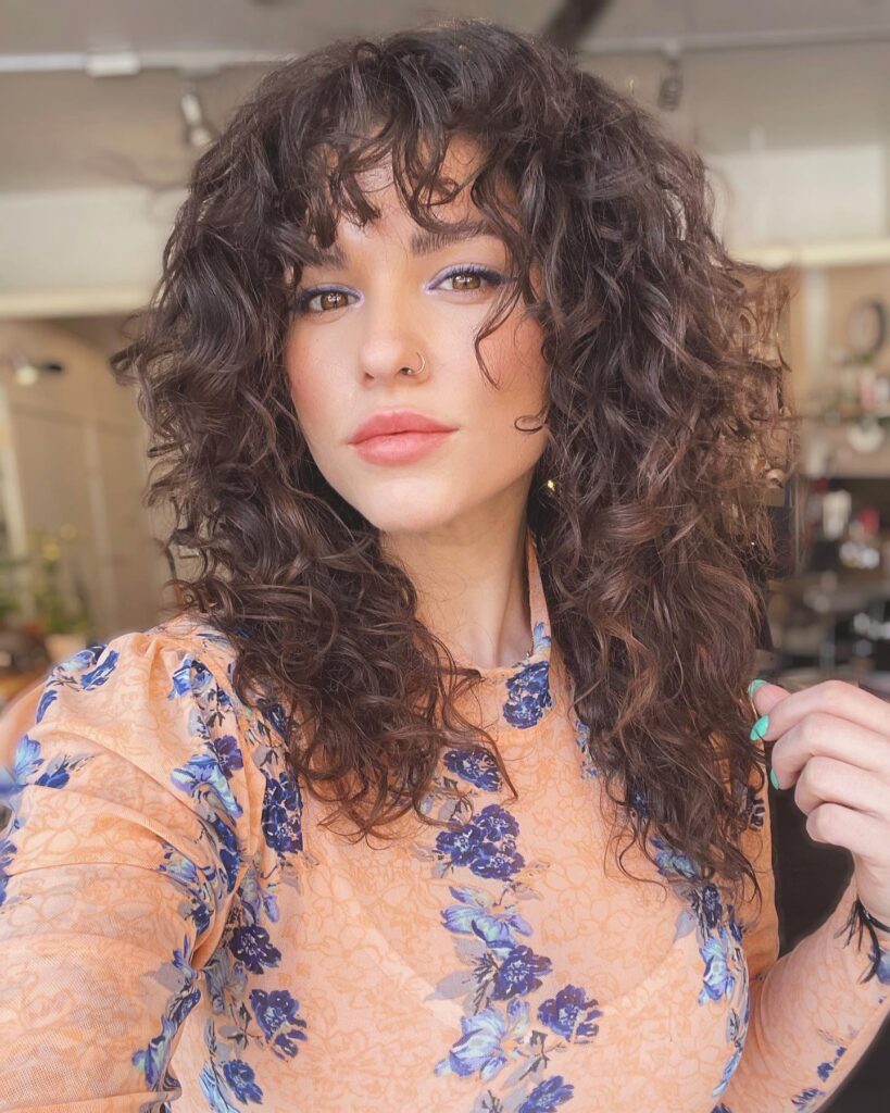 A woman with medium-length voluminous brown curly hair and curly bangs.
