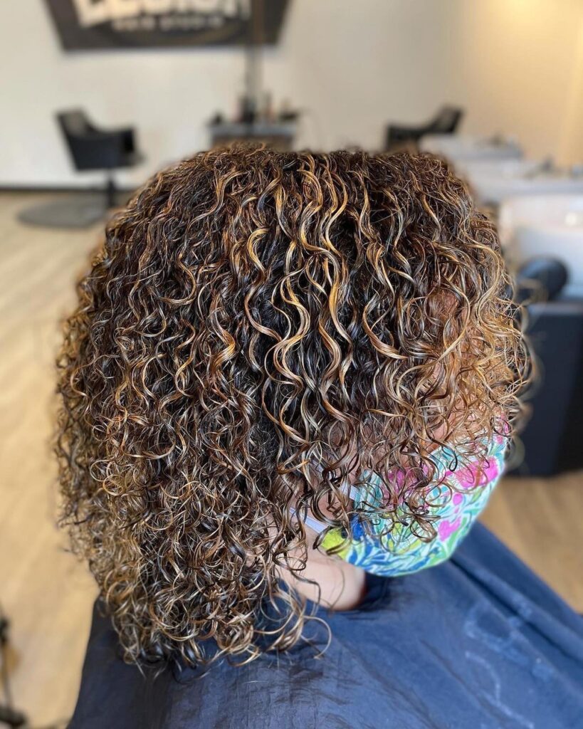 A person with shoulder-length curly hair featuring thin caramel highlights and highly defined ringlets.