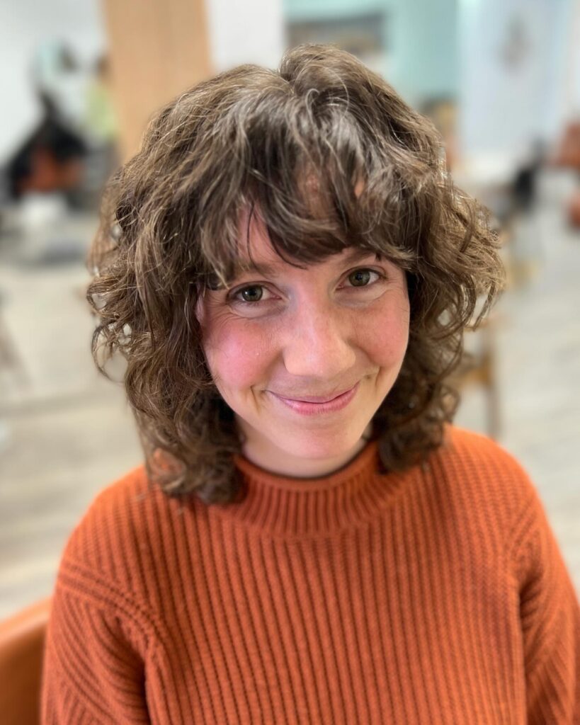 A smiling woman with a shoulder-length chestnut brown wavy shag hairstyle featuring soft layers and a full curly fringe.