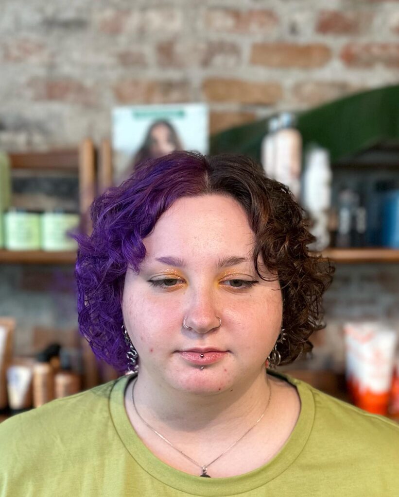 A person with an ear-length curly shag hairstyle featuring a split-color design of natural dark brown and bright purple.