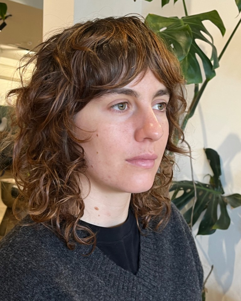 A woman with shoulder-length caramel brown wavy hair in a layered shag cut with feathered bangs.