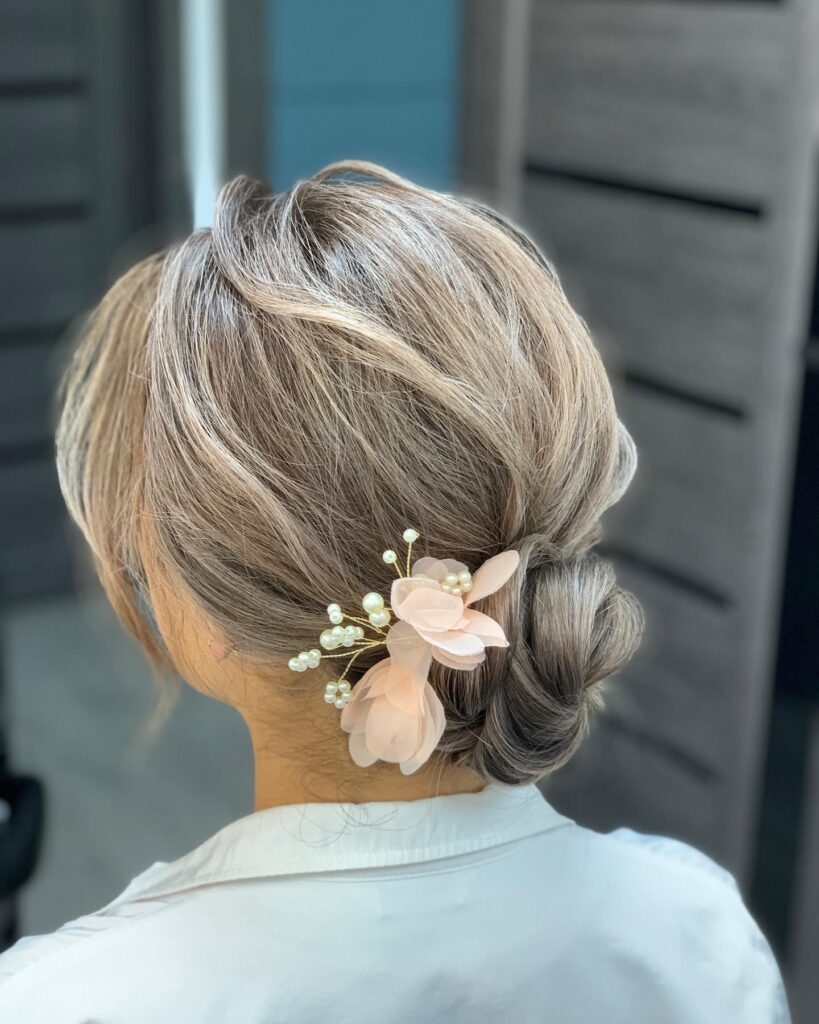 A woman with ash-blonde balayage hair has her locks swept back into a voluminous low bun decorated with a pink flower and pearl hairpiece.