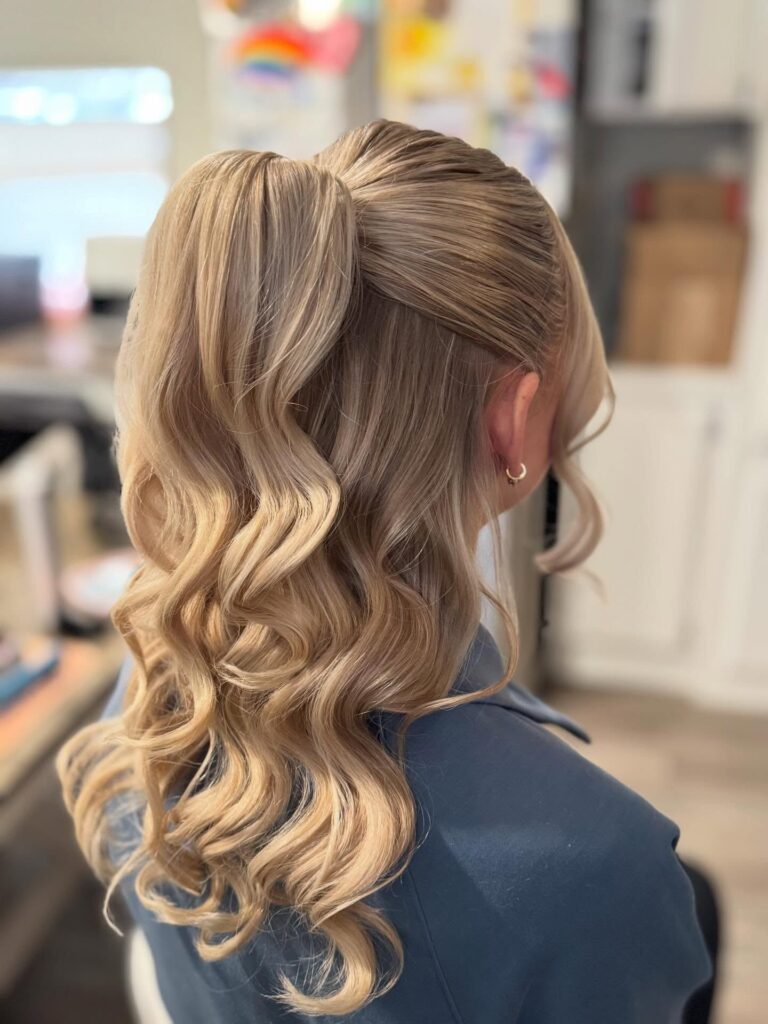 A woman with long, wavy blonde hair wears a high half-up ponytail with voluminous curls cascading down her back.