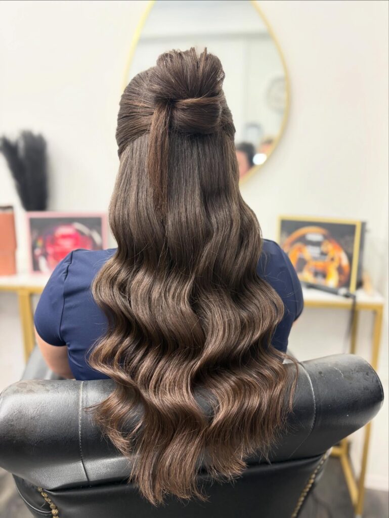 A woman with long, wavy, brunette hair wears a neat half-up topknot with the rest of her hair falling in smooth waves.