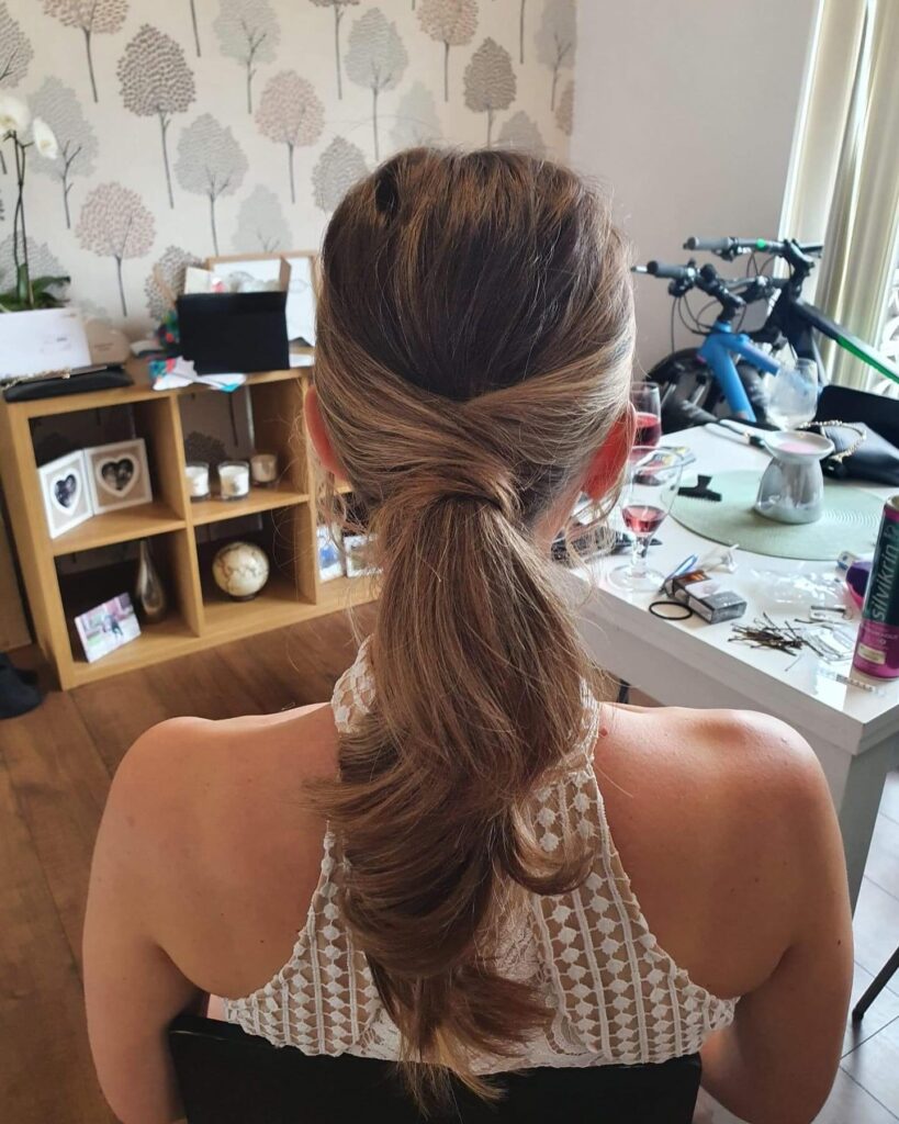A woman with medium-length highlighted brown hair wears a low ponytail with sections of hair crossed over the elastic.