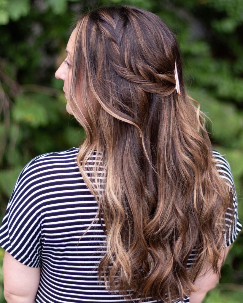 A woman with long, wavy, brown, and blonde-highlighted hair wears a thick side braid pulled back into a half-up style with a small clip.