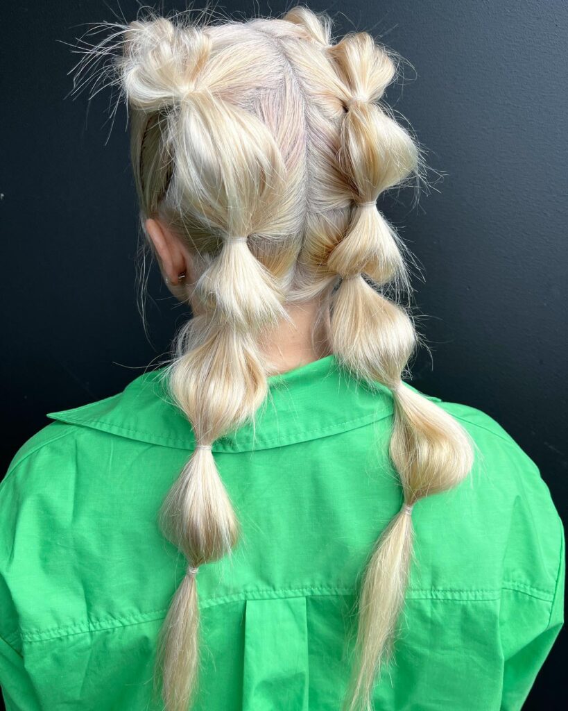 A woman with platinum blonde hair wears two pigtails styled into voluminous bubbles using clear elastic bands.