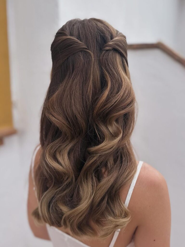 A woman with long, wavy, brown hair with caramel highlights wears a half-up style where the side sections are tucked and pinned at the back.