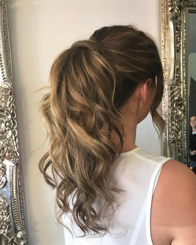 A woman with highlighted brown hair wears a voluminous, messy high ponytail with loose, wavy strands framing her face.