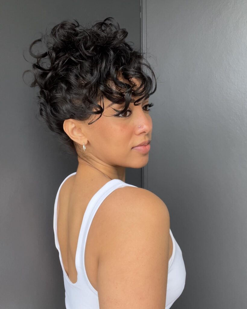 A woman with a dark curly faux hawk hairstyle featuring voluminous curls pinned up and falling forward over her forehead.