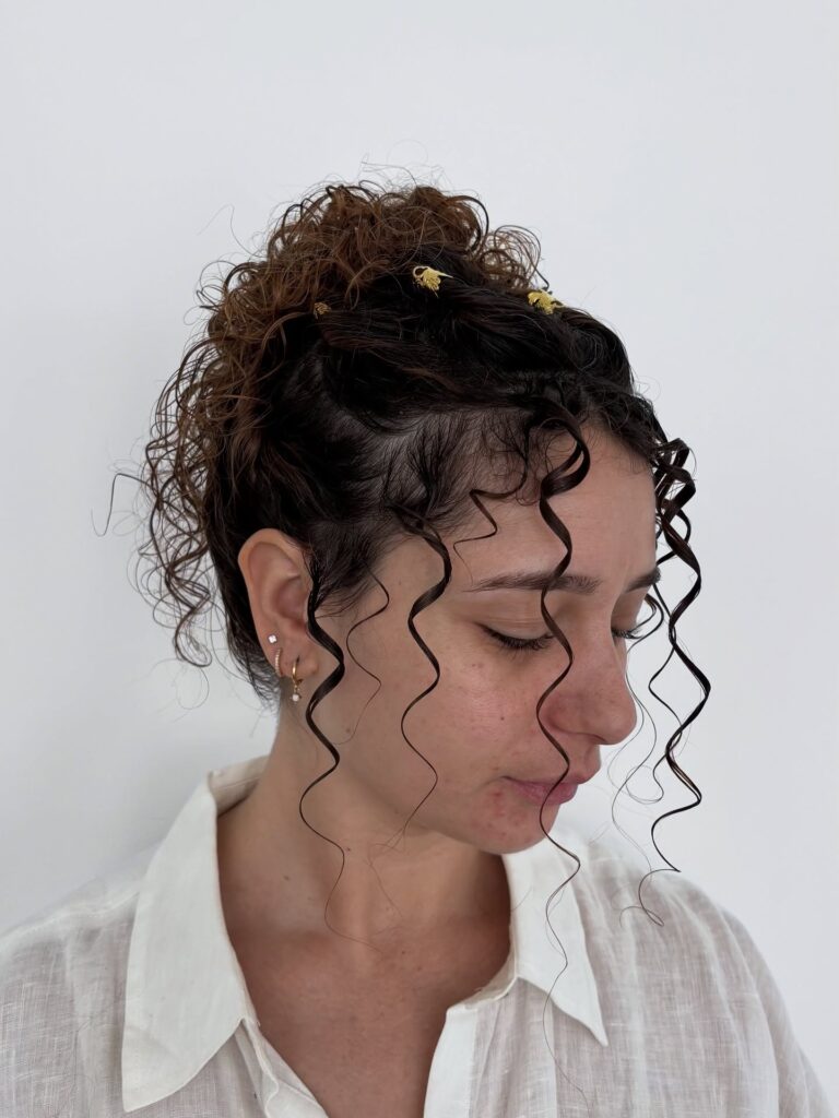 A woman with her curly hair in a high bun decorated with gold clips and long loose curls framing her face.