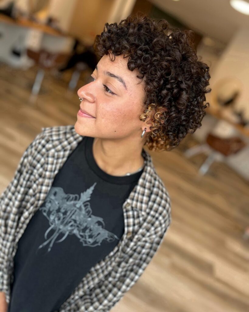 A woman with a short, tapered, curly haircut in dark brown featuring a small section of blonde curls near the ear.