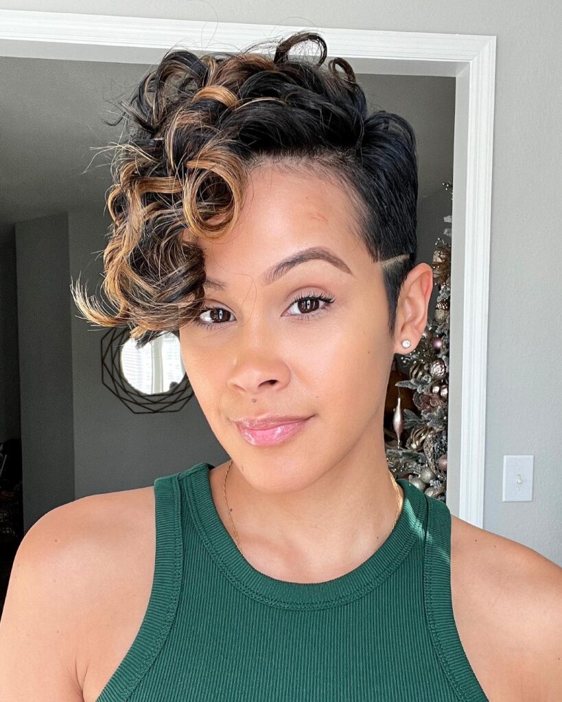 A woman with a short edgy pixie cut featuring a side-shaved line and long curly caramel-highlighted bangs swept to one side.