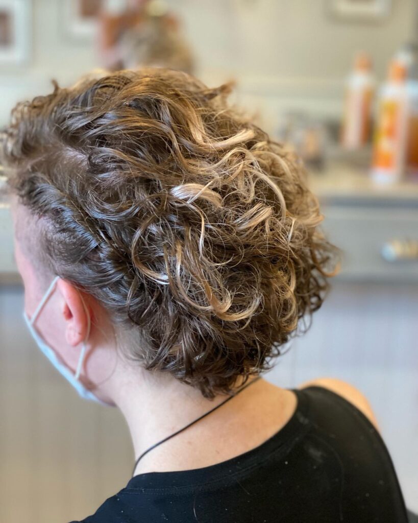 A back view of a woman with a short, tapered, curly haircut featuring sandy blonde highlights and voluminous layers on top.
