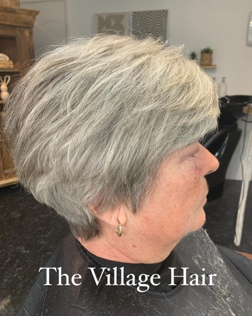 A side profile of a woman with a short textured ash grey pixie cut featuring soft layers and a tapered back.