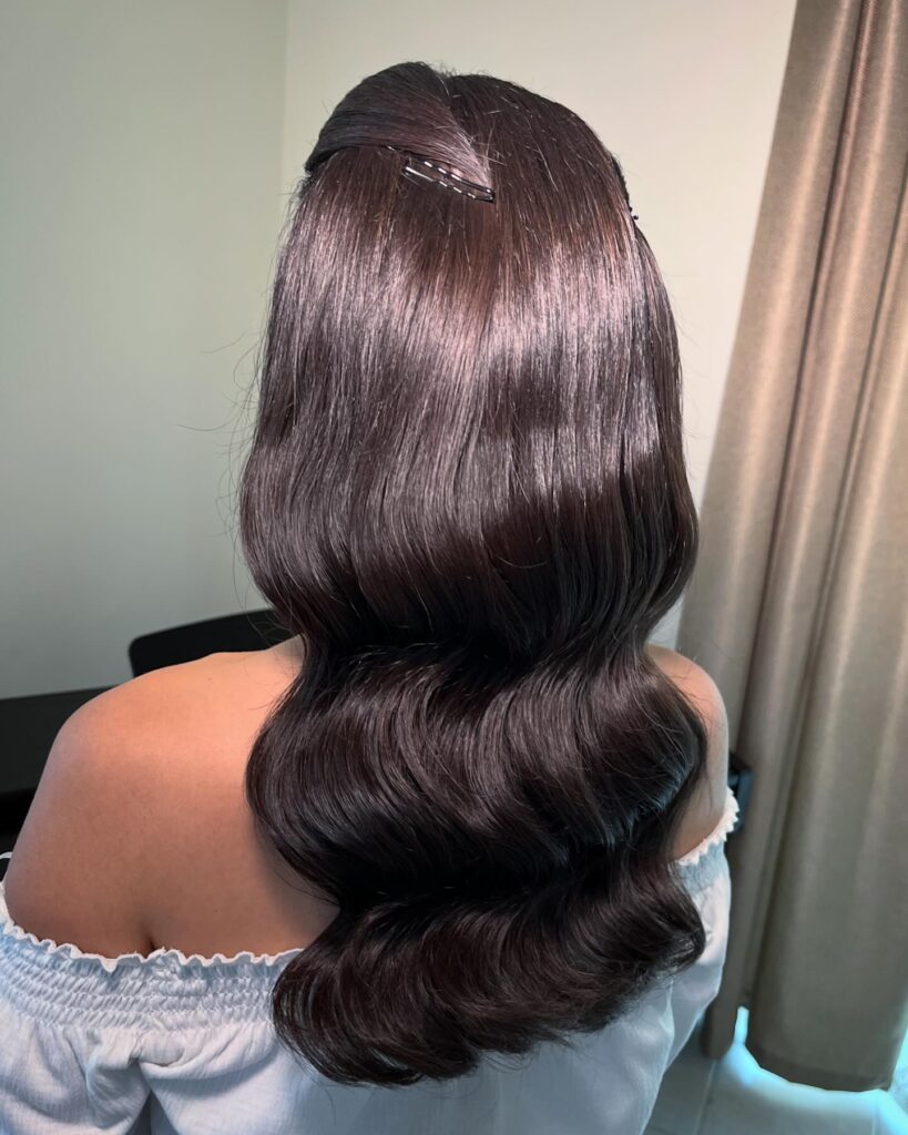 A woman from the back with long, glossy dark brown hair styled in wide vintage waves and pinned in a half-up style.